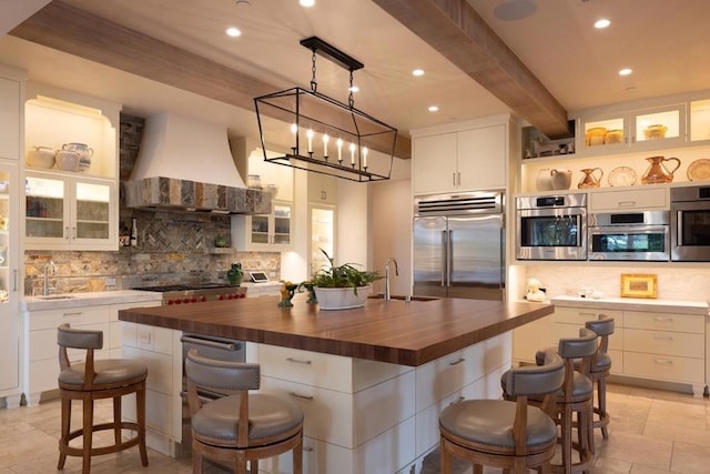 kitchen with butcher block counters, an island with sink, stainless steel appliances, custom range hood, and sink