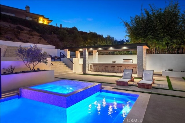 pool at dusk with an in ground hot tub, a patio area, and a bar