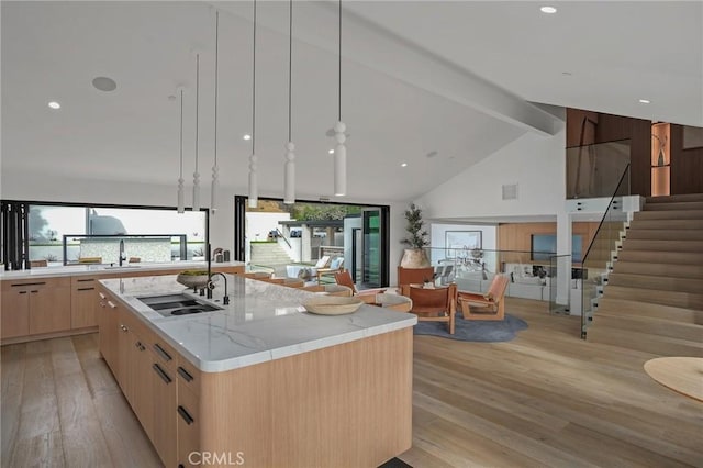 kitchen with an island with sink, beamed ceiling, light brown cabinetry, light stone countertops, and sink