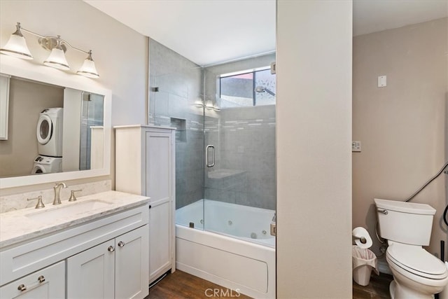 full bathroom featuring wood-type flooring, shower / bath combination with glass door, toilet, stacked washer and dryer, and vanity