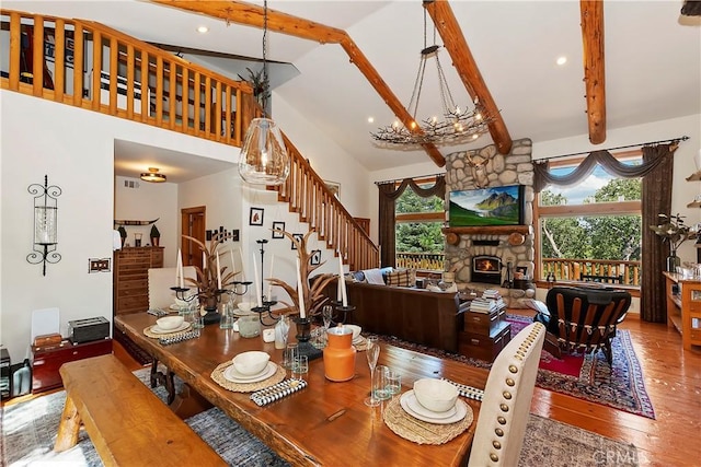 dining space with a high ceiling, wood-type flooring, beamed ceiling, a stone fireplace, and a chandelier