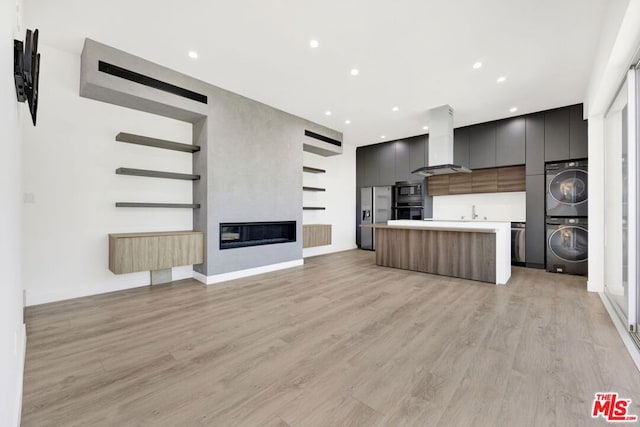 kitchen featuring a kitchen island, stacked washer / dryer, stainless steel fridge with ice dispenser, wall chimney range hood, and light wood-type flooring