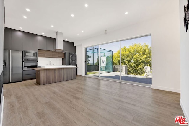 kitchen featuring a kitchen island, wall chimney exhaust hood, light hardwood / wood-style flooring, stacked washing maching and dryer, and appliances with stainless steel finishes