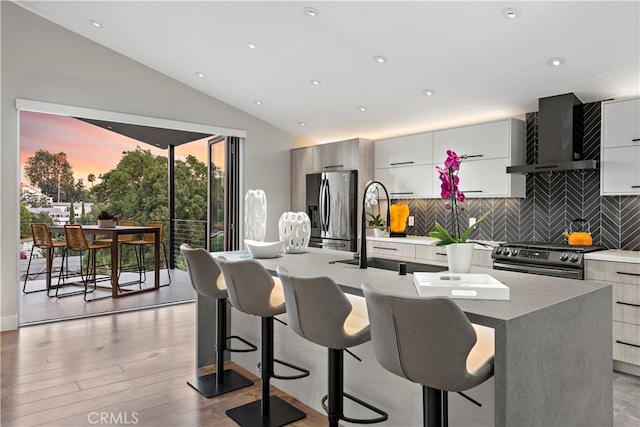 kitchen featuring an island with sink, wall chimney range hood, stainless steel appliances, a breakfast bar area, and decorative backsplash