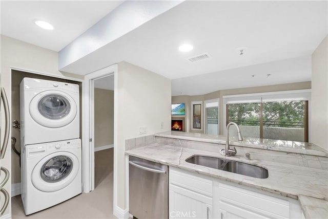 kitchen with light stone countertops, dishwasher, white cabinetry, stacked washer and dryer, and sink