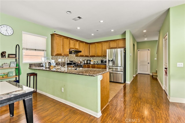 kitchen with kitchen peninsula, backsplash, stainless steel appliances, dark hardwood / wood-style floors, and dark stone countertops