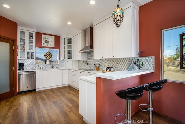 kitchen featuring dark wood-type flooring, wall chimney range hood, white cabinetry, dishwasher, and a kitchen bar