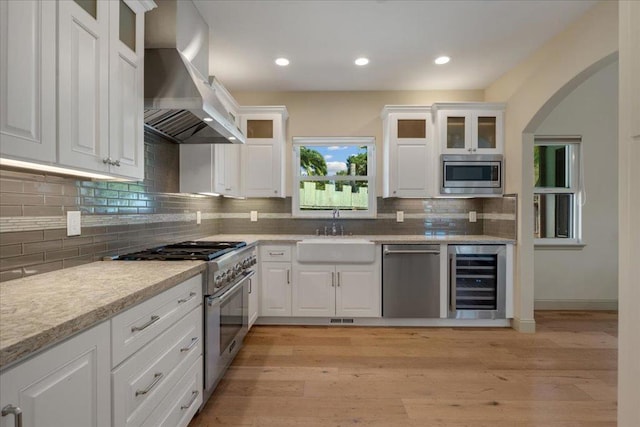 kitchen with wall chimney range hood, stainless steel appliances, white cabinets, wine cooler, and light hardwood / wood-style flooring