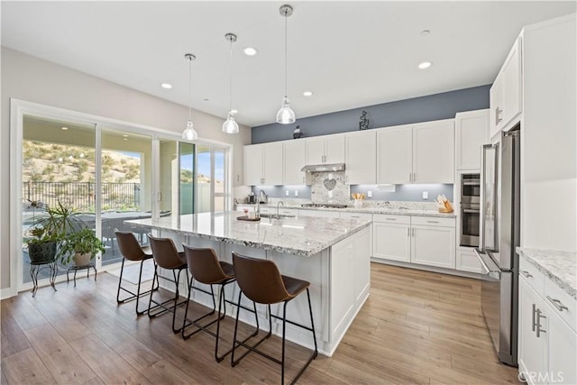 kitchen with hanging light fixtures, appliances with stainless steel finishes, white cabinets, light stone counters, and a kitchen island with sink