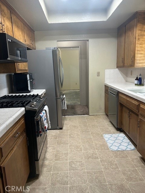 kitchen with appliances with stainless steel finishes, tasteful backsplash, a raised ceiling, tile counters, and sink