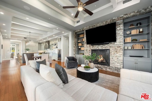 living room with a stone fireplace, wood-type flooring, beam ceiling, ceiling fan, and built in shelves