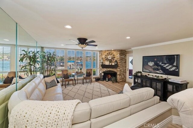 living room with crown molding, a stone fireplace, ceiling fan, and light wood-type flooring