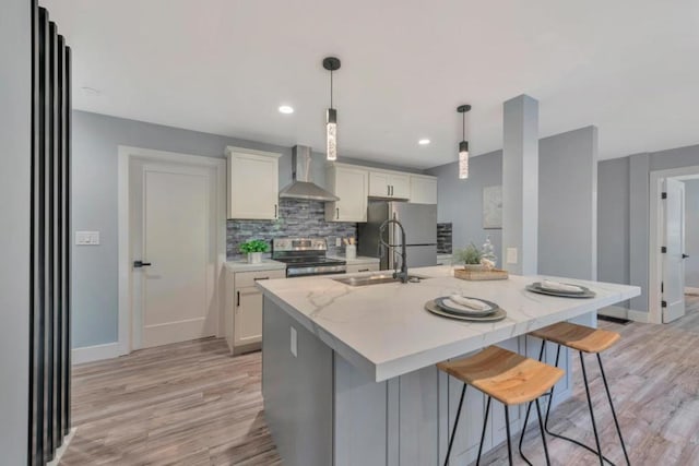 kitchen featuring light stone countertops, pendant lighting, appliances with stainless steel finishes, wall chimney range hood, and a kitchen island with sink