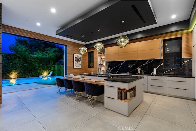 kitchen with a raised ceiling, hanging light fixtures, a kitchen island with sink, backsplash, and white cabinetry