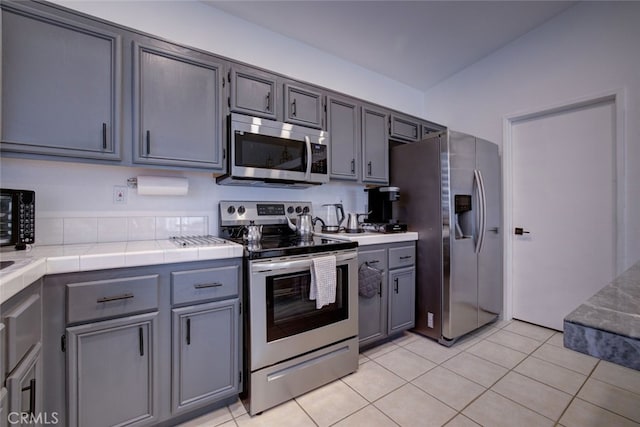 kitchen with light tile patterned floors, gray cabinets, stainless steel appliances, tile countertops, and lofted ceiling