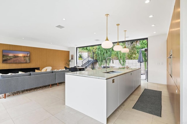 kitchen with a wall of windows, hanging light fixtures, white cabinets, a center island with sink, and sink