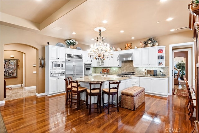 kitchen with white cabinetry, dark wood-type flooring, wall chimney exhaust hood, built in appliances, and a center island with sink