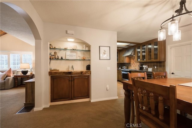 kitchen with wall chimney range hood, stainless steel stove, hanging light fixtures, backsplash, and dark carpet