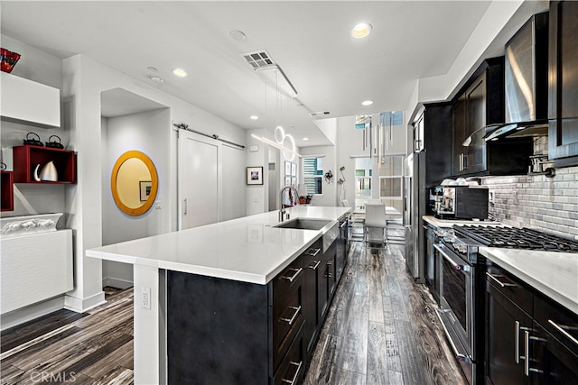 kitchen with dark wood-type flooring, hanging light fixtures, a barn door, a center island with sink, and appliances with stainless steel finishes