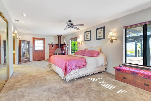 bedroom with multiple windows, light carpet, a wood stove, and ceiling fan