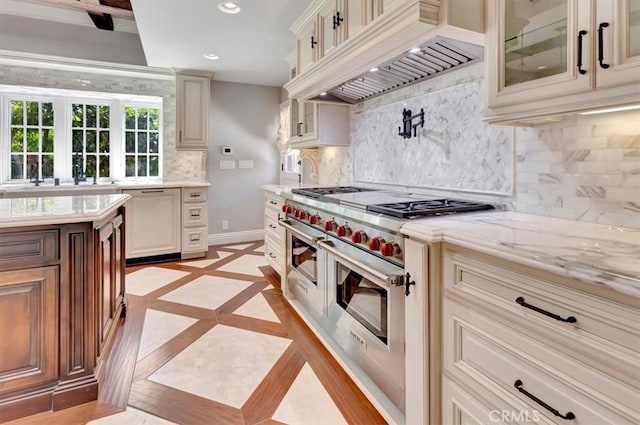 kitchen featuring premium range hood, range with two ovens, light stone counters, decorative backsplash, and cream cabinets