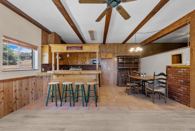 dining room with ceiling fan with notable chandelier, wooden walls, beam ceiling, and light hardwood / wood-style floors