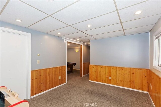 carpeted empty room with a drop ceiling, wood walls, and billiards