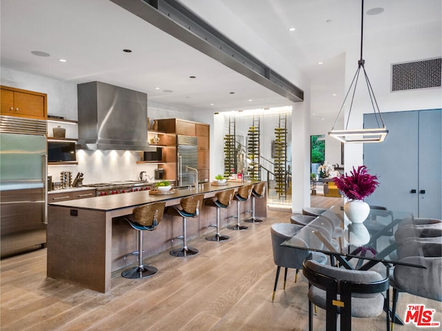 kitchen with stainless steel built in refrigerator, pendant lighting, a kitchen island with sink, wall chimney exhaust hood, and light hardwood / wood-style flooring