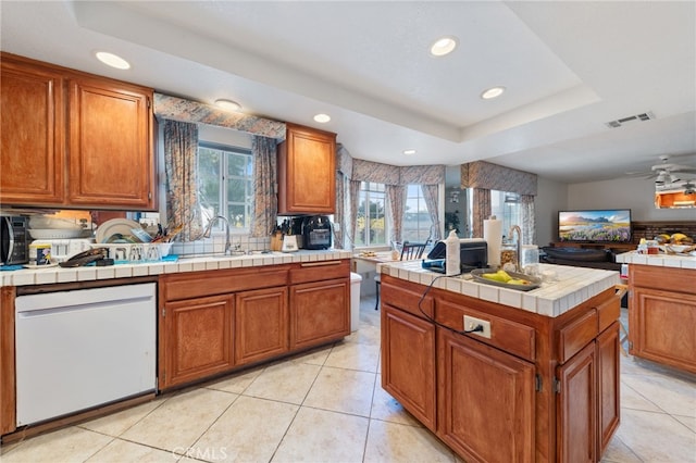 kitchen with ceiling fan, dishwasher, tile counters, sink, and a tray ceiling