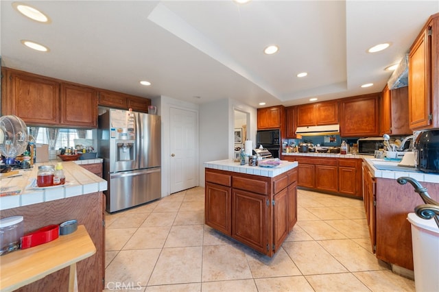 kitchen featuring tile countertops, a center island, black appliances, light tile patterned floors, and a tray ceiling