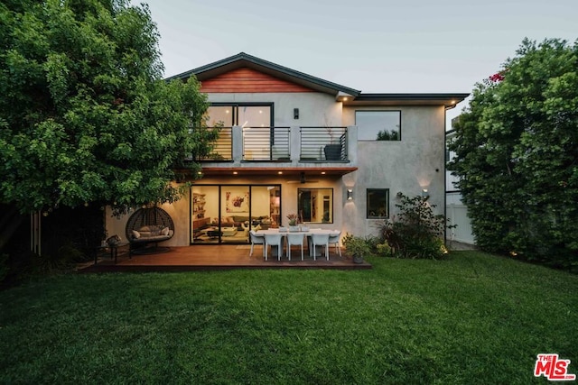 back house at dusk featuring a balcony, a patio area, and a yard