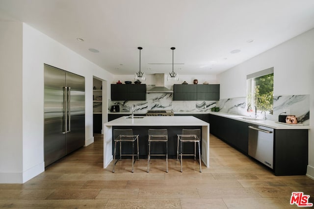 kitchen featuring a kitchen breakfast bar, hanging light fixtures, stainless steel appliances, a kitchen island with sink, and wall chimney range hood