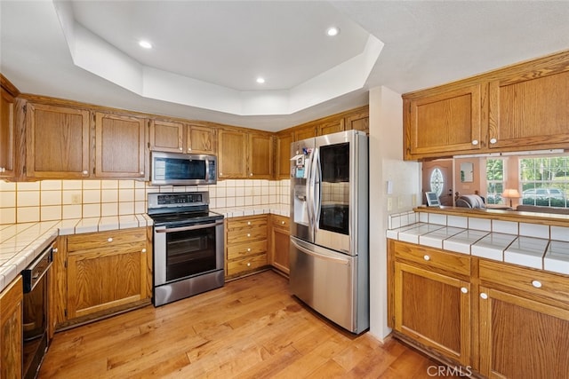 kitchen with light wood-type flooring, backsplash, a raised ceiling, appliances with stainless steel finishes, and tile countertops