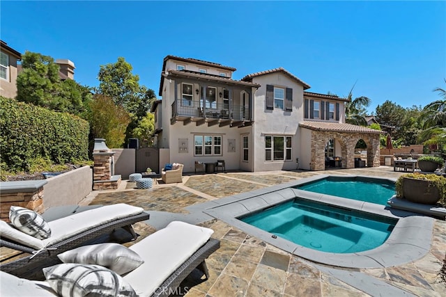 rear view of property with a patio, a balcony, exterior fireplace, and a pool with hot tub