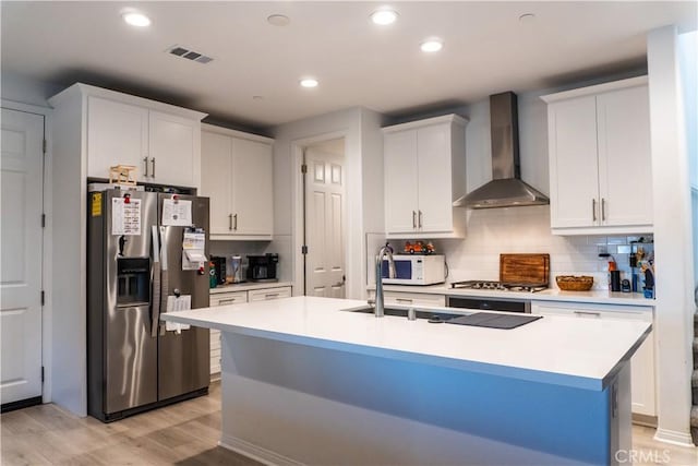 kitchen featuring a kitchen island with sink, white cabinets, wall chimney exhaust hood, light wood-type flooring, and appliances with stainless steel finishes