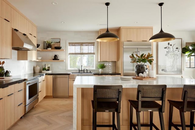 kitchen with light brown cabinetry, stainless steel appliances, hanging light fixtures, and light parquet floors