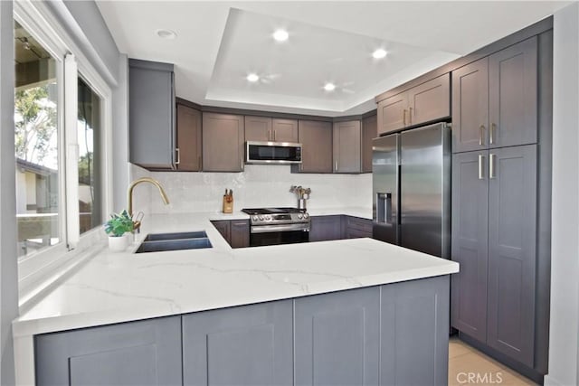 kitchen featuring sink, a raised ceiling, kitchen peninsula, stainless steel appliances, and light stone counters