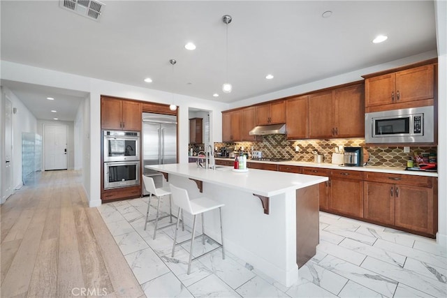 kitchen with a breakfast bar, pendant lighting, built in appliances, a center island with sink, and light hardwood / wood-style floors