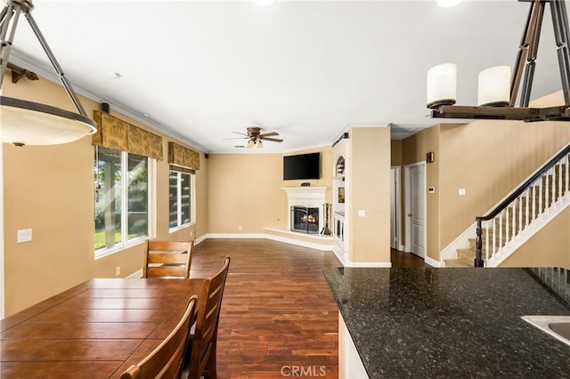 unfurnished dining area with dark wood-style flooring, baseboards, a warm lit fireplace, stairway, and ceiling fan