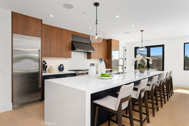 kitchen with an island with sink, hanging light fixtures, wall chimney range hood, stainless steel appliances, and light hardwood / wood-style floors