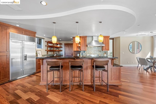 kitchen with pendant lighting, hardwood / wood-style floors, wall chimney range hood, and built in fridge