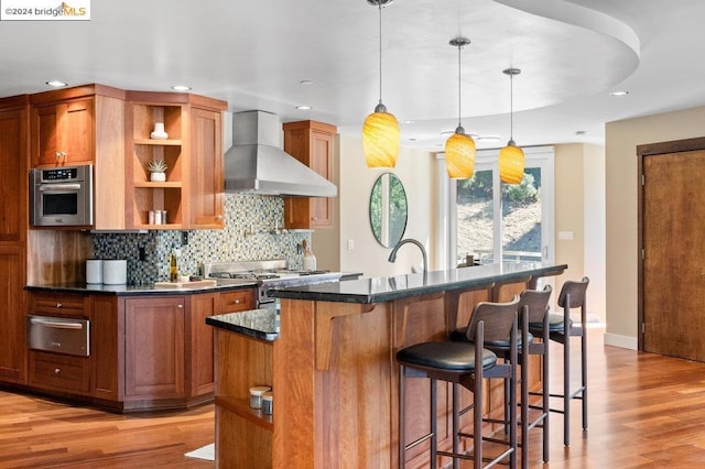 kitchen featuring an island with sink, decorative light fixtures, wall chimney range hood, appliances with stainless steel finishes, and light wood-type flooring