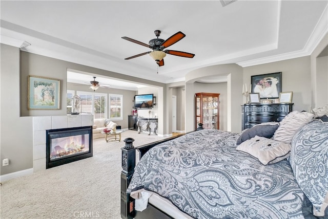 carpeted bedroom with a tray ceiling, crown molding, a tile fireplace, and ceiling fan