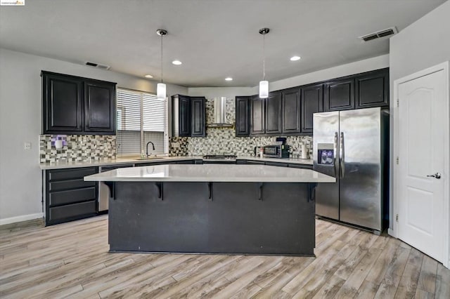 kitchen featuring light wood-type flooring, wall chimney exhaust hood, stainless steel appliances, pendant lighting, and a center island