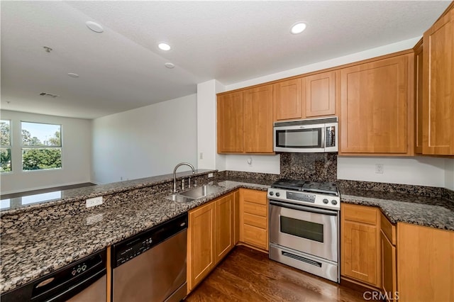 kitchen with appliances with stainless steel finishes, kitchen peninsula, dark hardwood / wood-style floors, dark stone counters, and sink