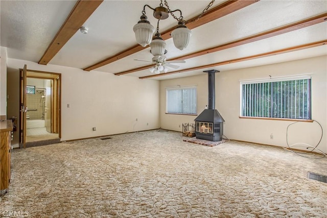 unfurnished living room with carpet flooring, ceiling fan, beam ceiling, and a wood stove