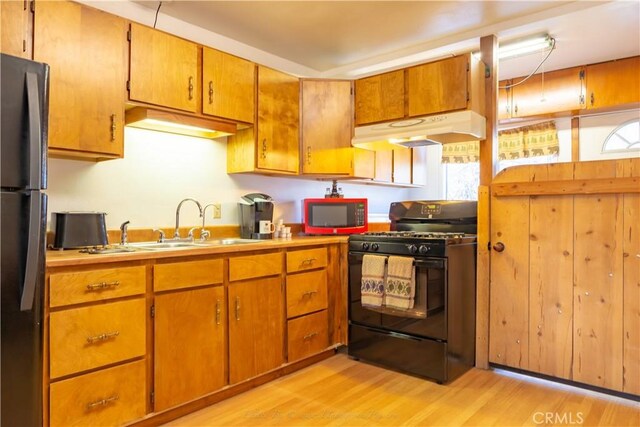 kitchen with brown cabinets, black appliances, under cabinet range hood, a sink, and light wood finished floors