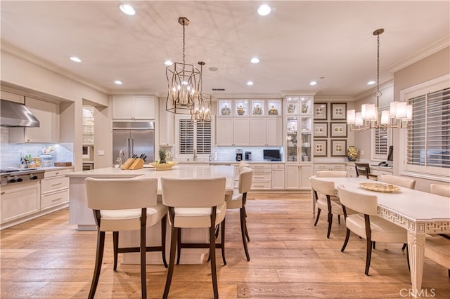 kitchen featuring a large island with sink, wall chimney exhaust hood, pendant lighting, stainless steel appliances, and light hardwood / wood-style flooring