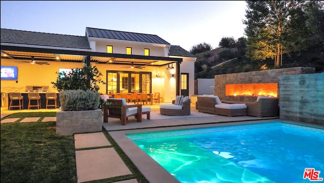 pool at dusk with an outdoor living space with a fireplace, a bar, ceiling fan, and a patio area