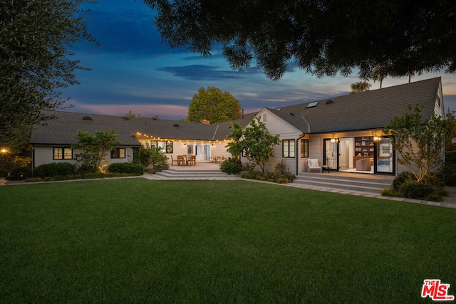 back house at dusk featuring a patio and a lawn